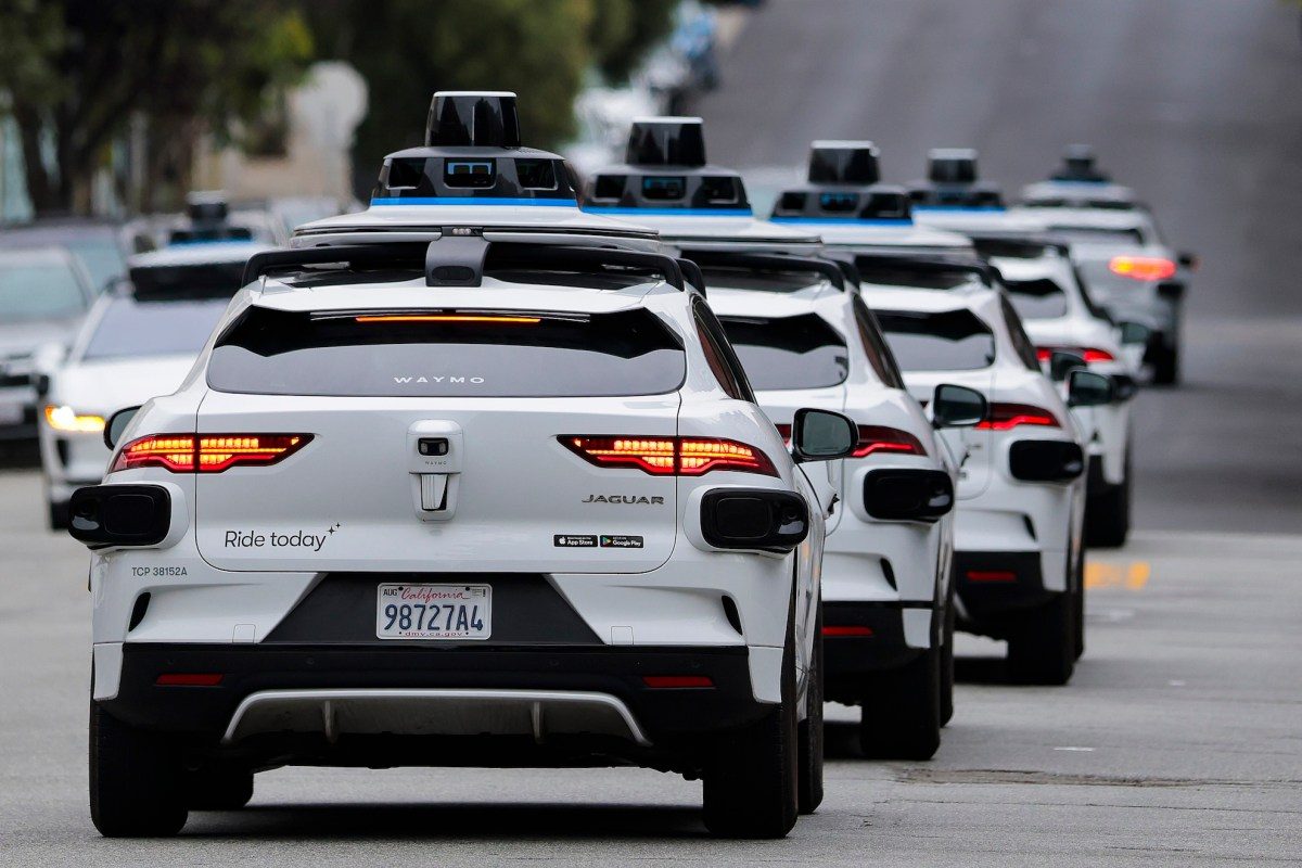 A Waymo self-driving car stopped on a city street with emergency lights in the distance