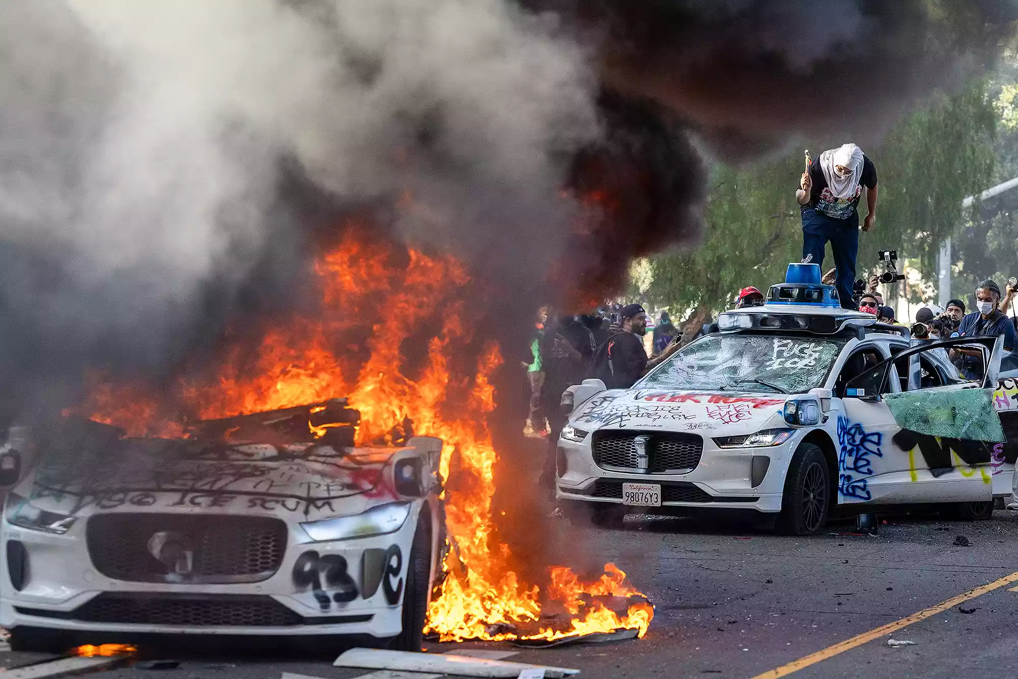 TOPSHOT - Demonstrators smash the windshield of a vehicle next to a burning Waymo vehicle as protesters clash with law enforcement in the streets surrounding the federal building during a protest following federal immigration operations in Los Angeles, California, on June 8, 2025. Demonstrators torched cars and scuffled with security forces in Los Angeles on June 8, as police kept protestors away from the National Guard troops President Donald Trump sent to the streets of the second biggest US city